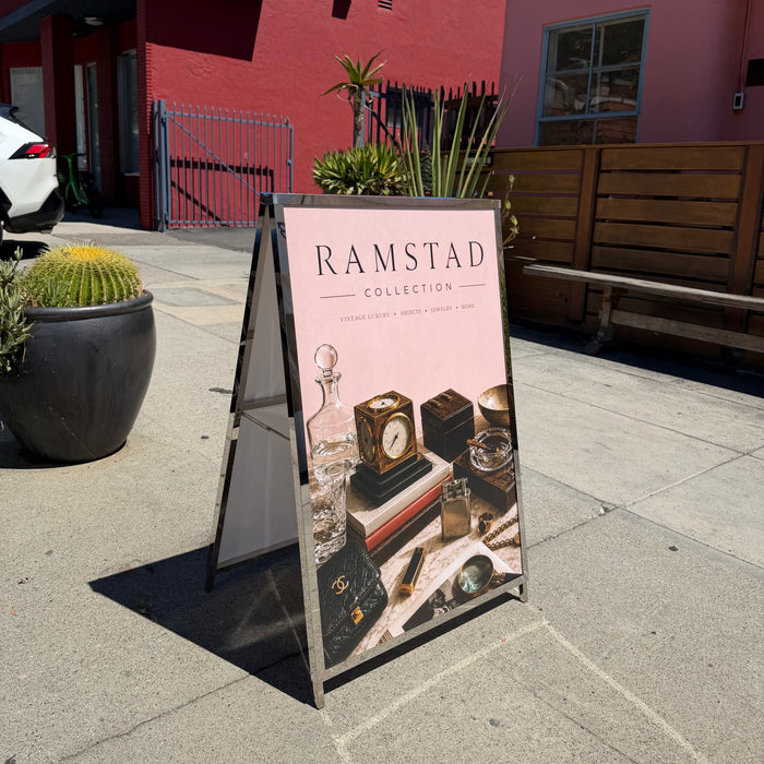 Metal A-frame sign holder displaying a pink Ramstad poster outside a red building.