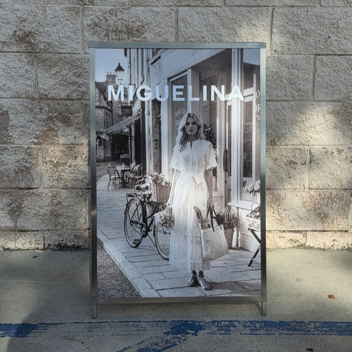 Front view of an A-frame sign holder displaying a Miguelina fashion poster.