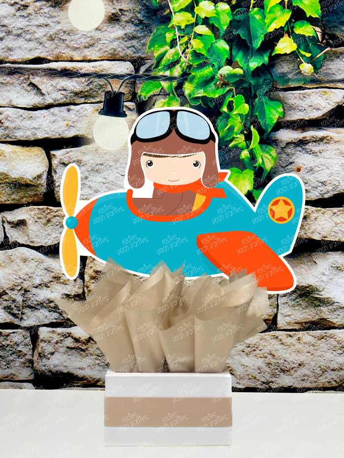 Set of boy airplane pilot-themed birthday party table centerpieces from Rosie's Posh Parties. Features a personalized sign with orange wings, two boy pilots flying blue and orange airplanes, and a pilot flying a blue helicopter. The cutouts are nestled in square bases decorated with blue, tan, and brown tissue paper.