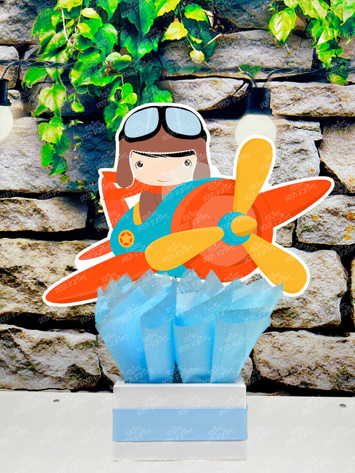 Set of boy airplane pilot-themed birthday party table centerpieces from Rosie's Posh Parties. Features a personalized sign with orange wings, two boy pilots flying blue and orange airplanes, and a pilot flying a blue helicopter. The cutouts are nestled in square bases decorated with blue, tan, and brown tissue paper.