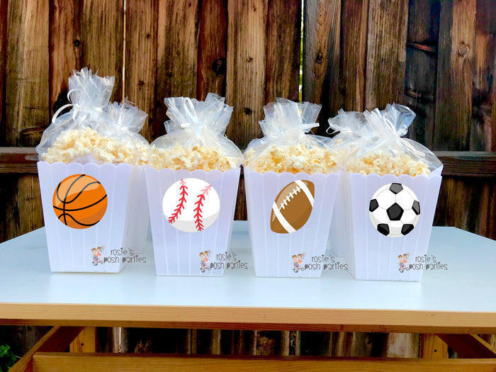 reusable white plastic popcorn bins Each individual bin features a different cartoon sports ball decal: a baseball, a soccer ball, a basketball, and a football. Inside each clear bin, a filled bag of popcorn is visible, tied at the top with a clear bag and ribbon. The bins have a classic scalloped edge design at the top