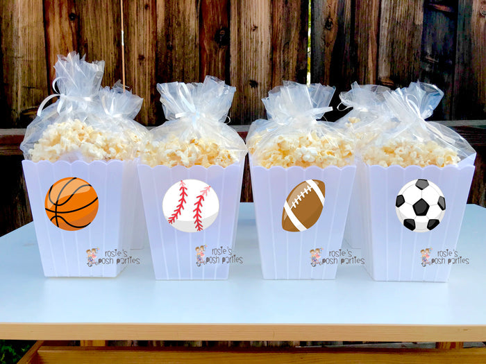 reusable white plastic popcorn bins Each individual bin features a different cartoon sports ball decal: a baseball, a soccer ball, a basketball, and a football. Inside each clear bin, a filled bag of popcorn is visible, tied at the top with a clear bag and ribbon. The bins have a classic scalloped edge design at the top