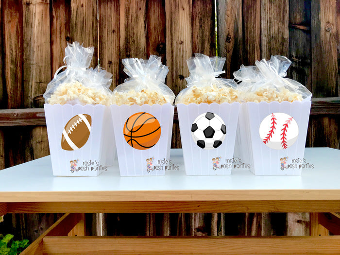 reusable white plastic popcorn bins Each individual bin features a different cartoon sports ball decal: a baseball, a soccer ball, a basketball, and a football. Inside each clear bin, a filled bag of popcorn is visible, tied at the top with a clear bag and ribbon. The bins have a classic scalloped edge design at the top