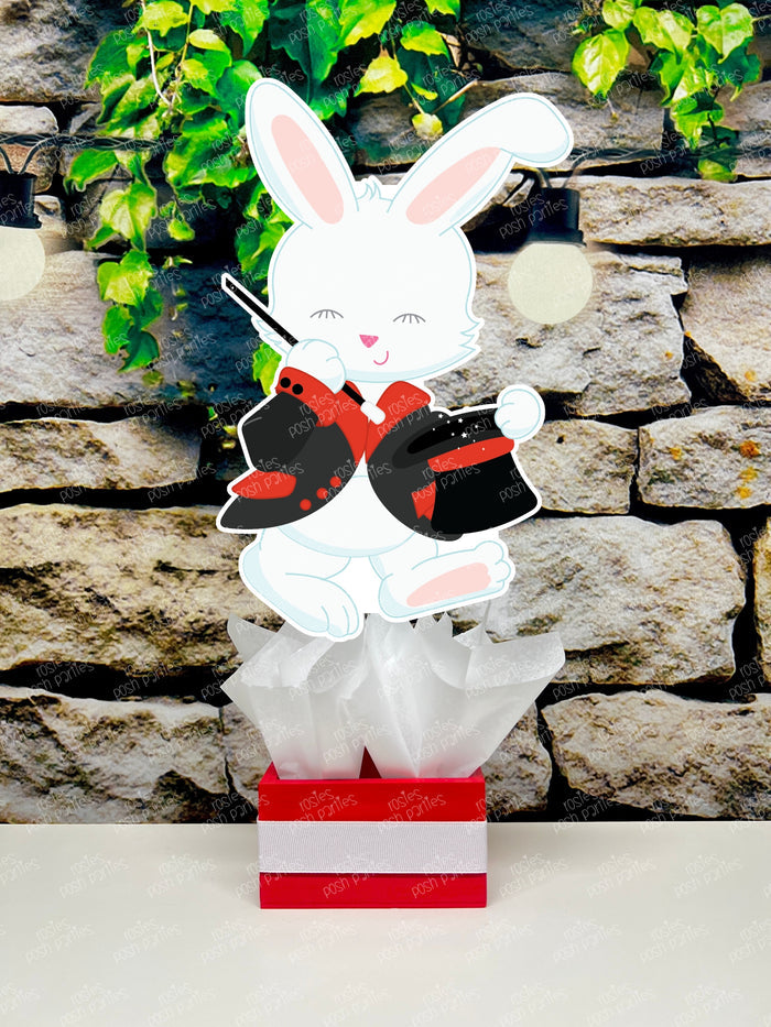 Kids birthday party magician themed centerpieces. The centerpieces feature illustrated cutouts including playing cards, a white rabbit in a tuxedo, a girl magician, a boy magician, a rabbit popping out of a top hat, and a magician's hat with a wand. Each cutout is set in a red, white, or black base filled with matching tissue paper. Receive fully assembled like pictured.