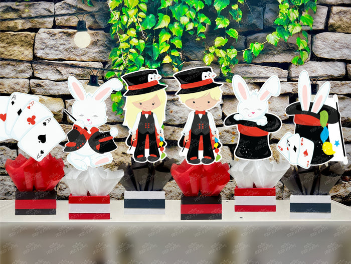 Kids birthday party magician themed centerpieces. The centerpieces feature illustrated cutouts including playing cards, a white rabbit in a tuxedo, a girl magician, a boy magician, a rabbit popping out of a top hat, and a magician's hat with a wand. Each cutout is set in a red, white, or black base filled with matching tissue paper. Receive fully assembled like pictured.