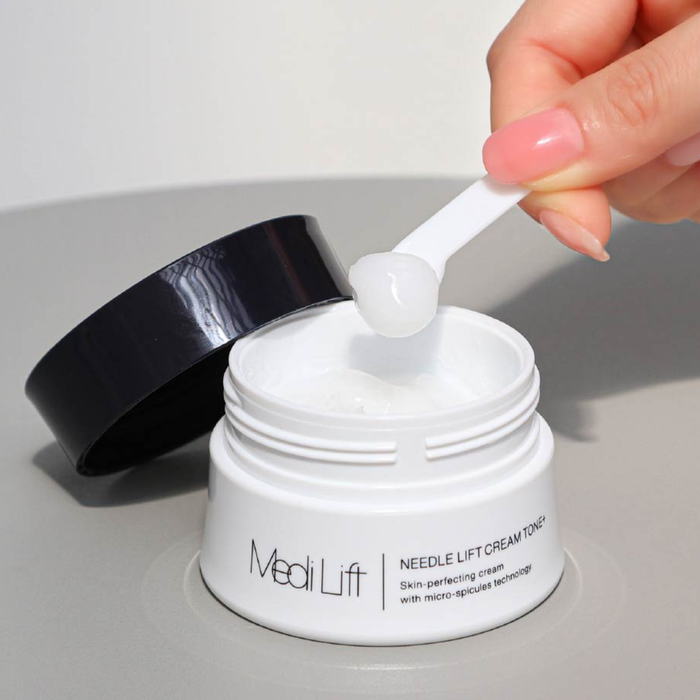 Hand using a white applicator to scoop cream from a MediLift jar on a light gray background