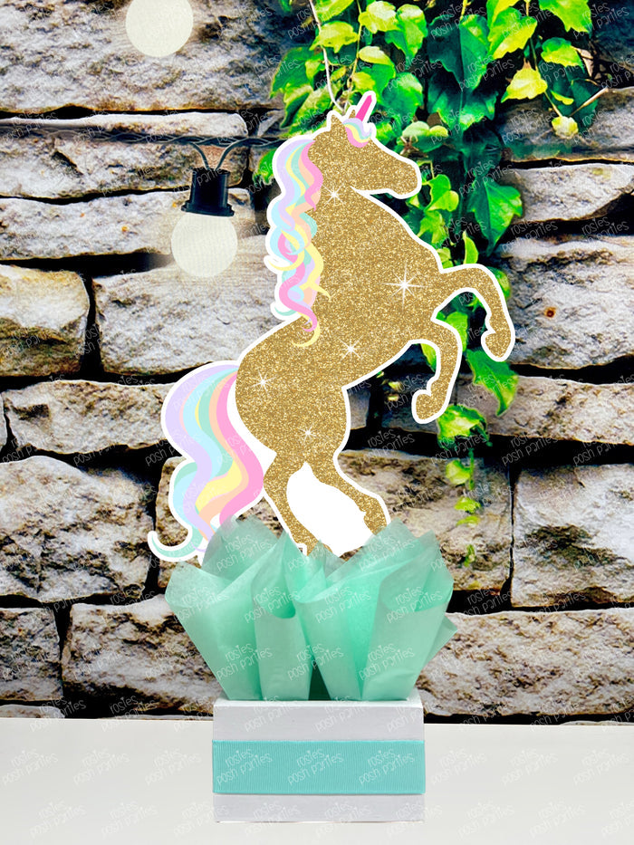 fully decorated unicorn theme table centerpieces for a birthday party or baby shower. Each centerpiece features a cute, pastel color unicorn with printed glitter accents, placed in a white box with colorful tissue paper in shades of blue, purple, mint green, and pink.