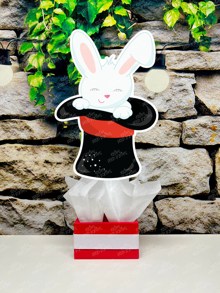 Kids birthday party magician themed centerpieces. The centerpieces feature illustrated cutouts including playing cards, a white rabbit in a tuxedo, a girl magician, a boy magician, a rabbit popping out of a top hat, and a magician's hat with a wand. Each cutout is set in a red, white, or black base filled with matching tissue paper. Receive fully assembled like pictured.