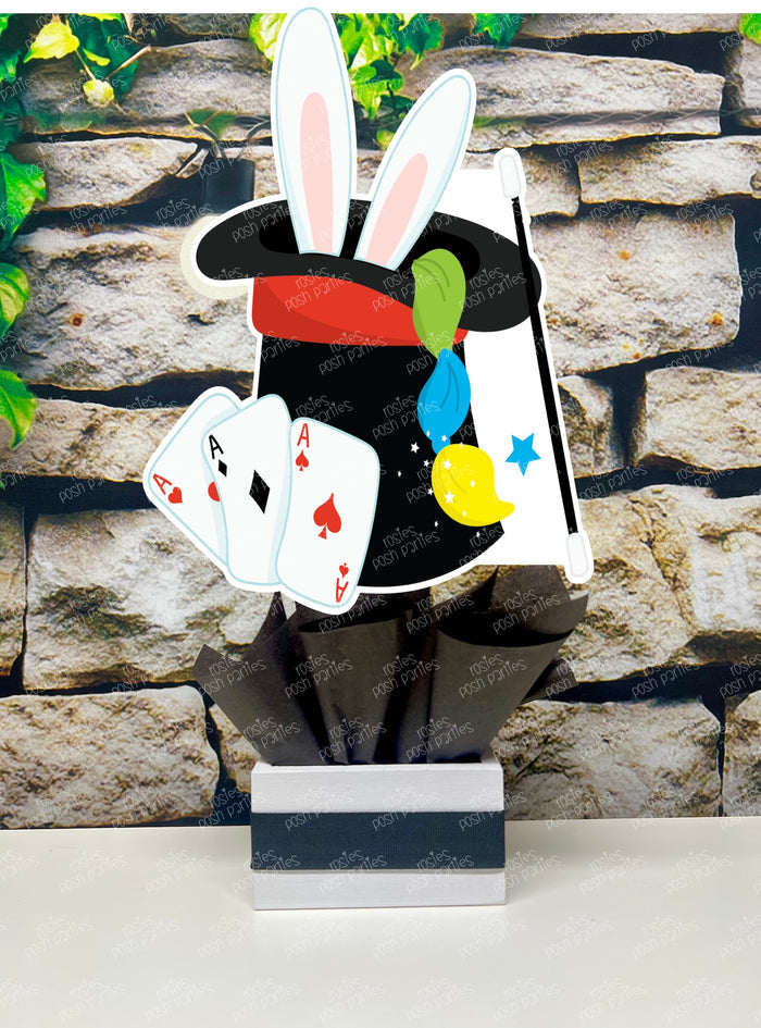 Kids birthday party magician themed centerpieces. The centerpieces feature illustrated cutouts including playing cards, a white rabbit in a tuxedo, a girl magician, a boy magician, a rabbit popping out of a top hat, and a magician's hat with a wand. Each cutout is set in a red, white, or black base filled with matching tissue paper. Receive fully assembled like pictured.