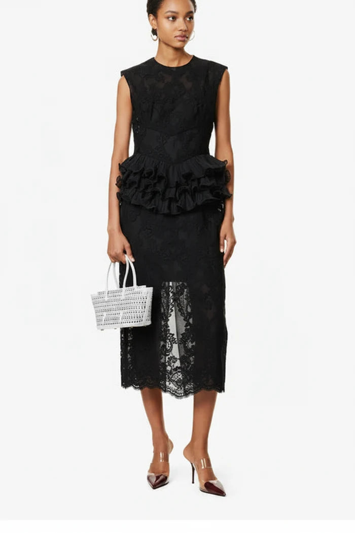 Woman wearing a black lace dress with ruffled details, holding a white woven handbag on a white background.
