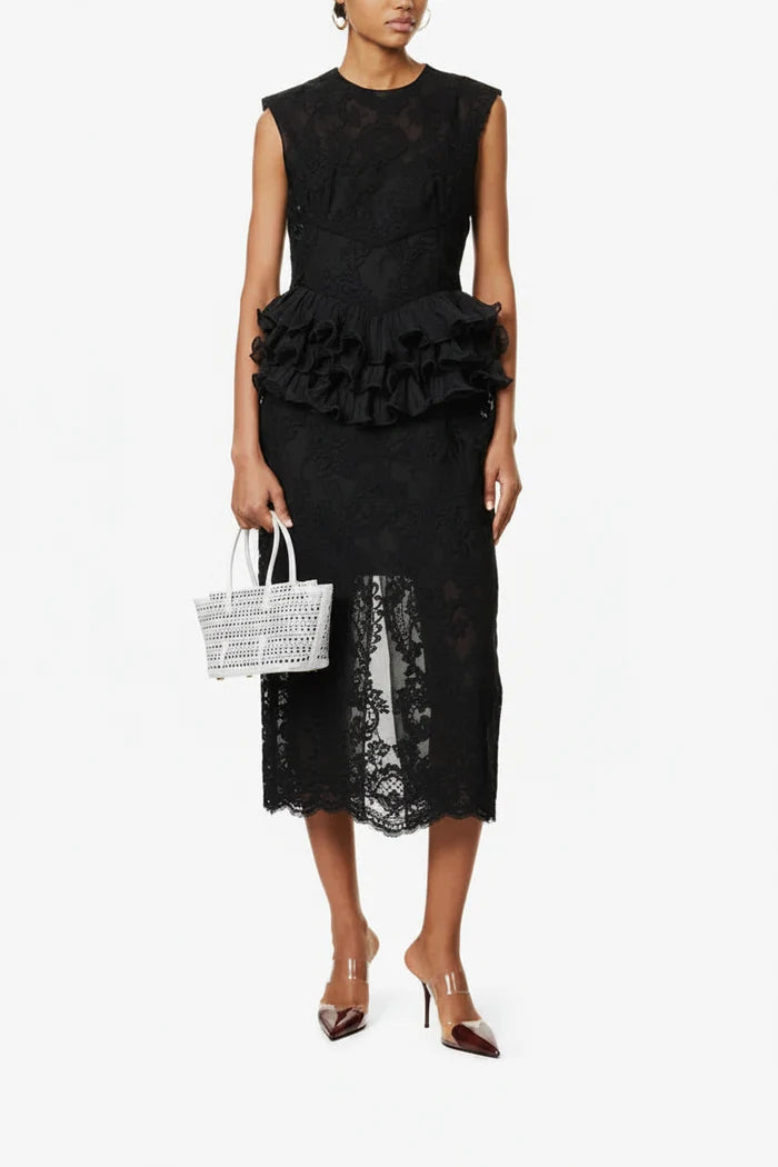 Woman wearing a black lace dress with ruffled details, holding a white woven handbag on a white background.