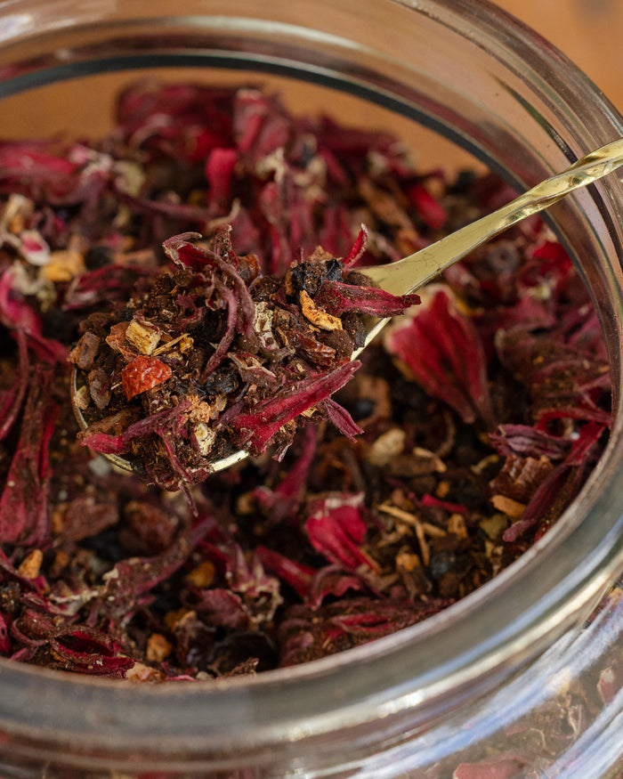 Close-up of a spoonful of dried herbs over a glass jar filled with similar herbs.